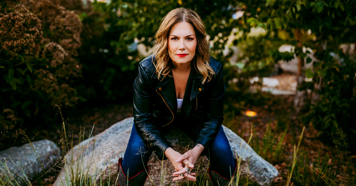 Portrait of Jane Kleeb leaning forward on a rock in a green landscape.